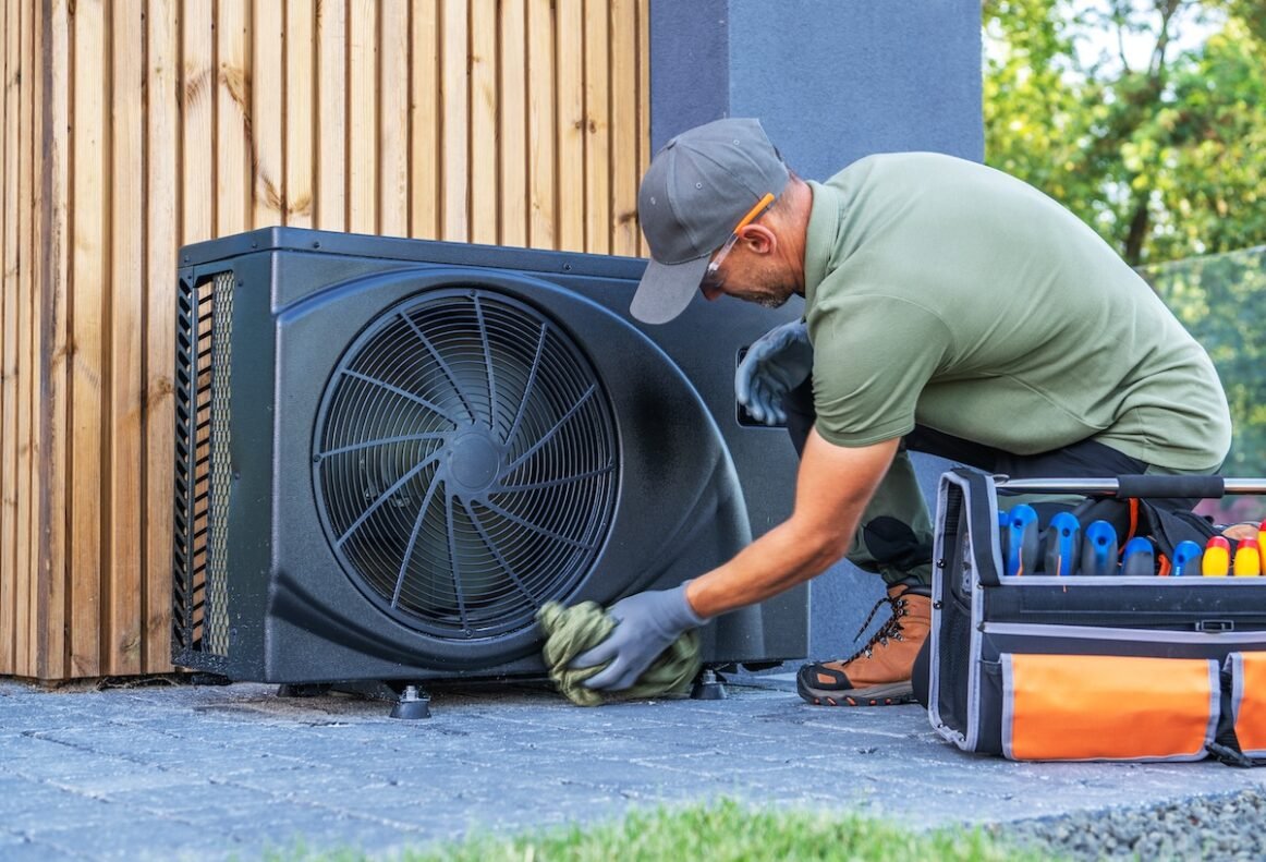 A technician in work attire cleans an outdoor air conditioning unit near a wooden wall. Tools are organized beside him for efficient service.