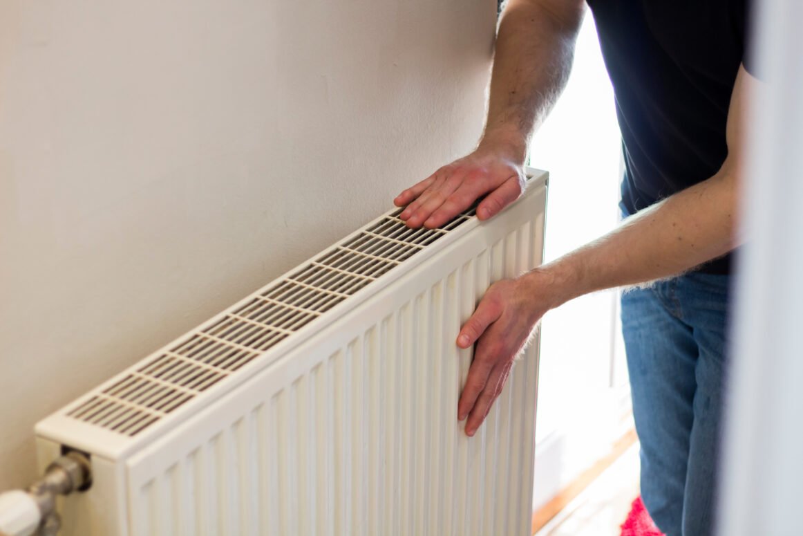 Man is checking radiator is it heating. Man warming his hands on the radiator. Home central heating system. Man is checking radiator is it heating. Man warming his hands on the radiator. Home central heating system.